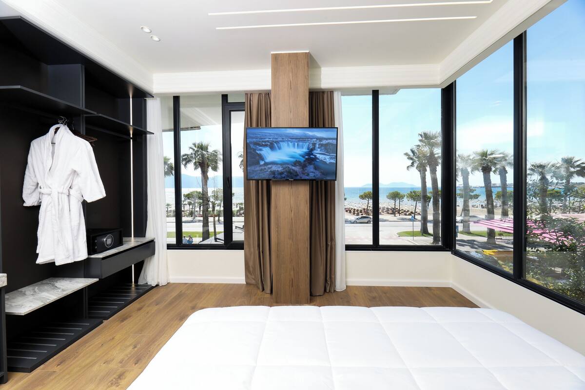 A modern hotel room with a large bed, a TV mounted on a wooden panel, a wardrobe, and a view of palm trees and a beach.