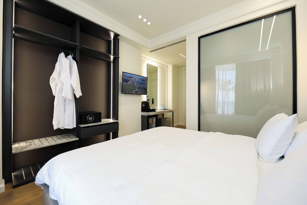 A modern hotel room with a large bed, a wardrobe, a desk with a computer, and a TV on the wall.