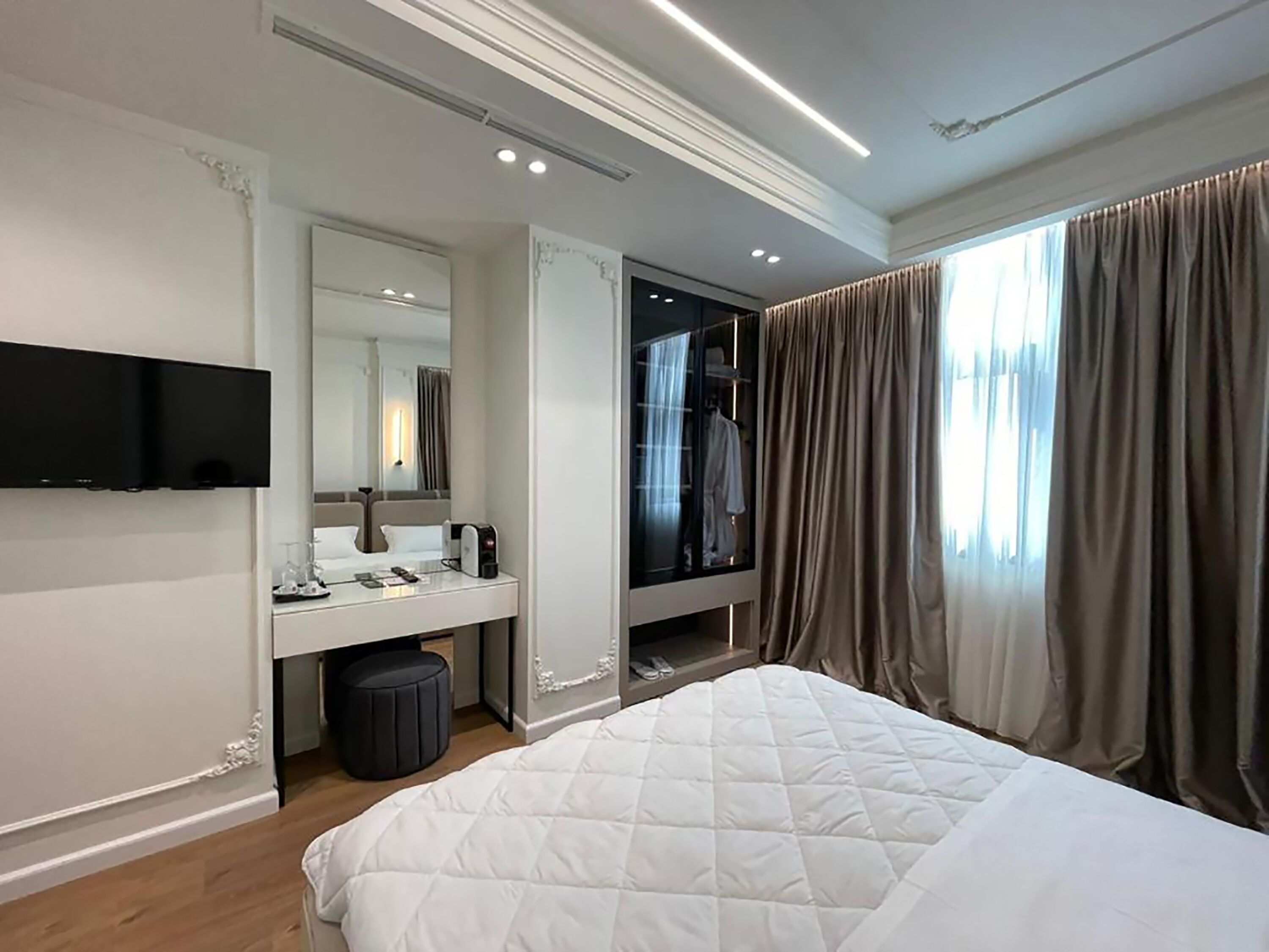 A modern hotel room with a large bed, a flat-screen TV mounted on the wall, a vanity with a mirror, and a walk-in closet.