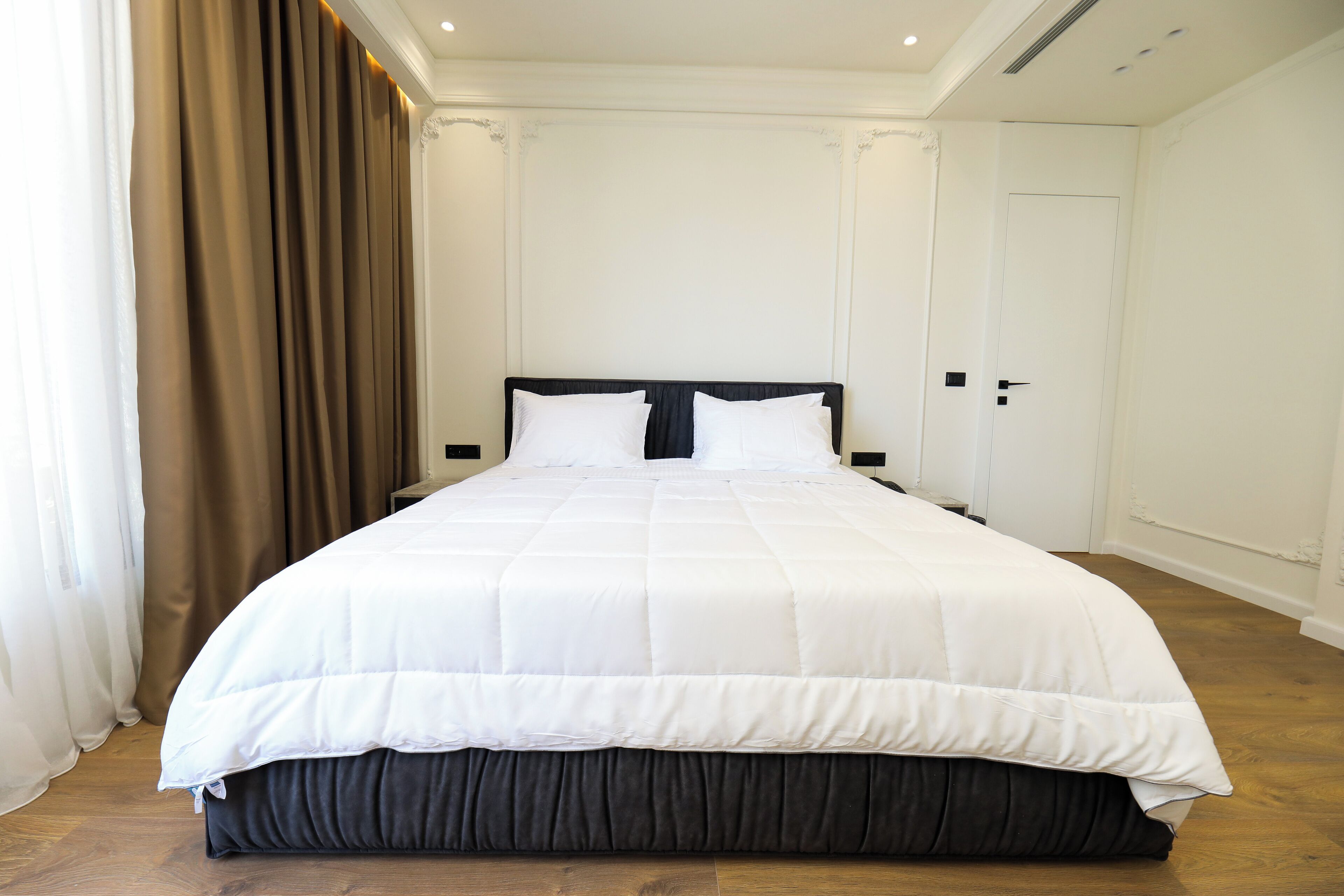 Una cama grande y bien hecha, con cabecera oscura y ropa de cama blanca, en una habitación con suelo de madera y paredes blancas.