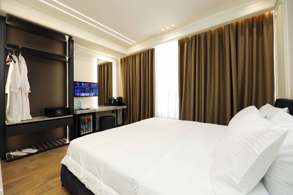 A hotel room with a large bed, a desk with a flat-screen TV, a minibar, and a wardrobe with hanging clothes.