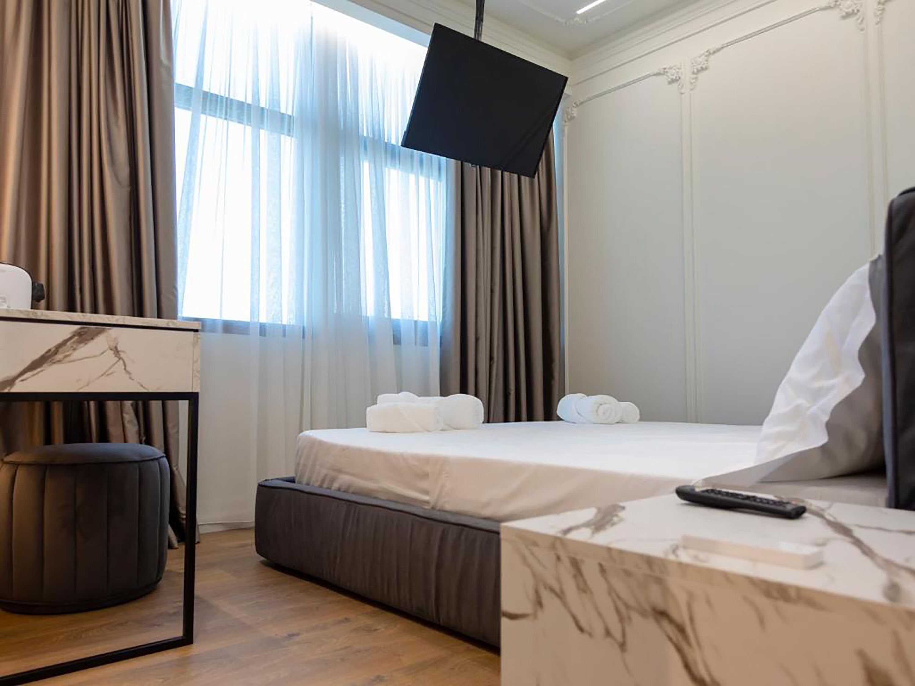 A hotel room with a bed, a desk with a chair, a small ottoman, and a large window with curtains.