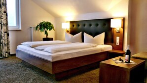 A hotel room with a large bed, two bedside lamps, a potted plant, a wooden nightstand, and a small table with a bottle and glass. - Weinhotel Kienle (Burrweiler)