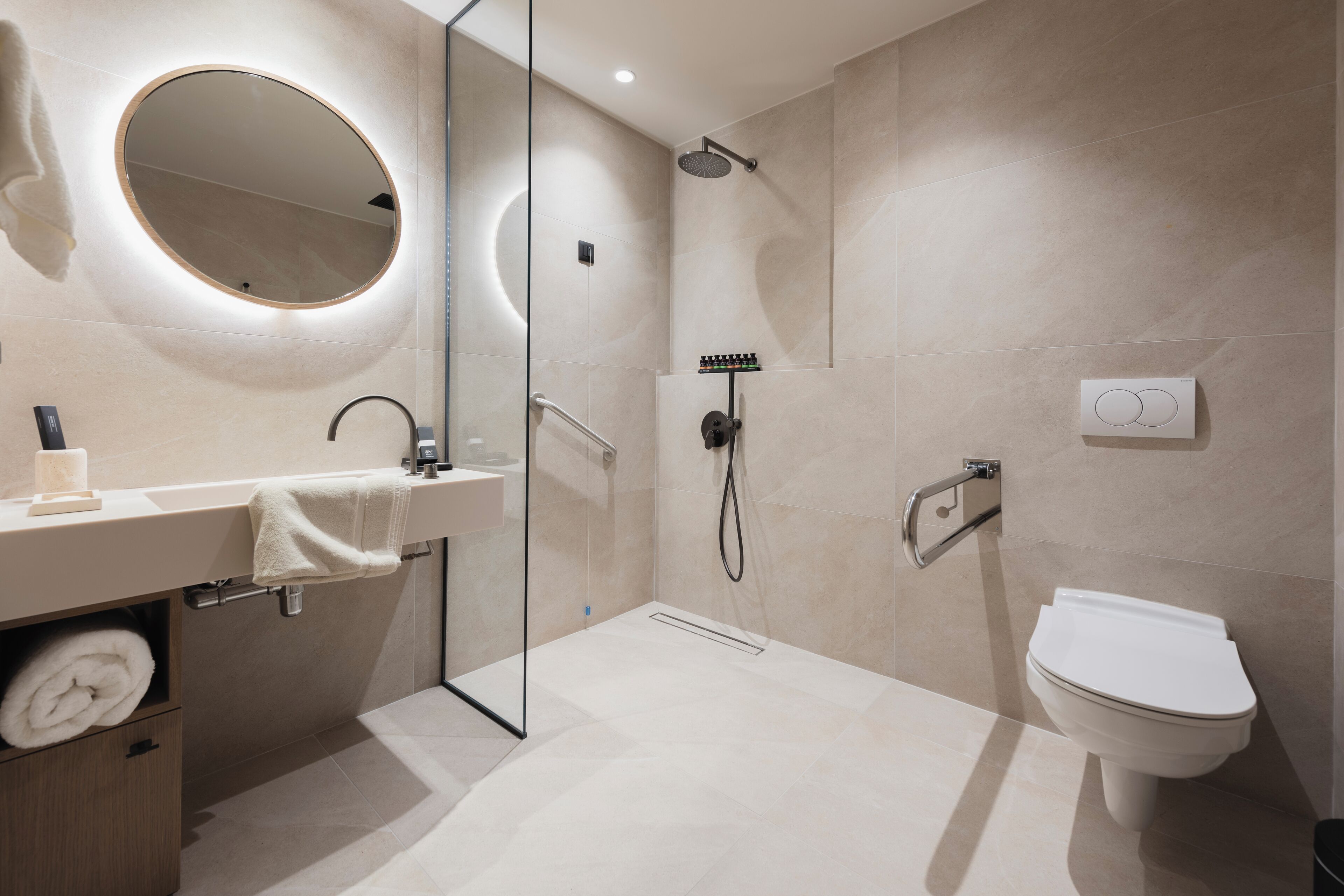 Une salle de bain moderne avec une douche à parois de verre, un miroir rond, des toilettes et un lavabo équipé d’un porte-serviettes.