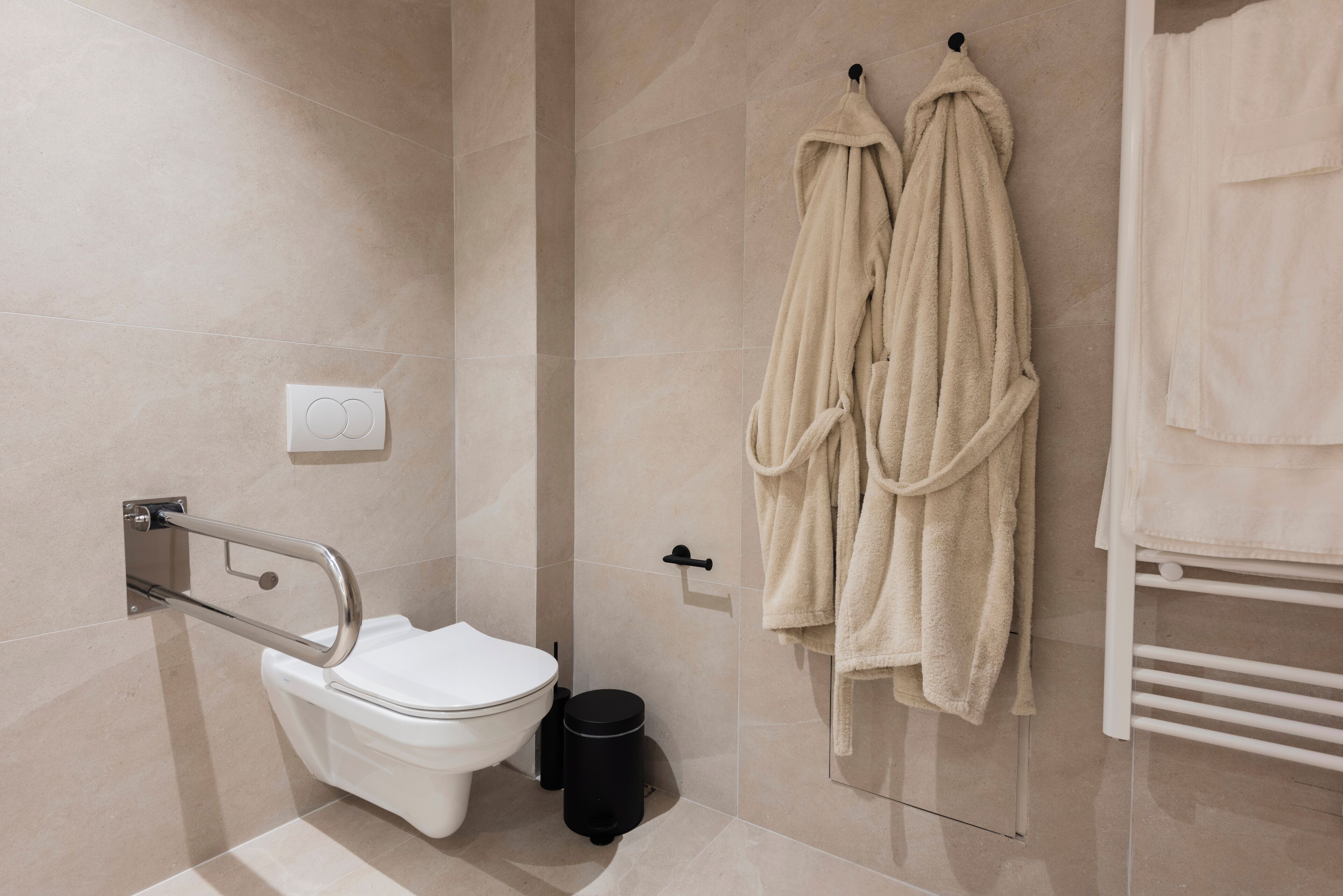A modern bathroom with a toilet, grab bars, a towel rack with robes, and a trash bin.