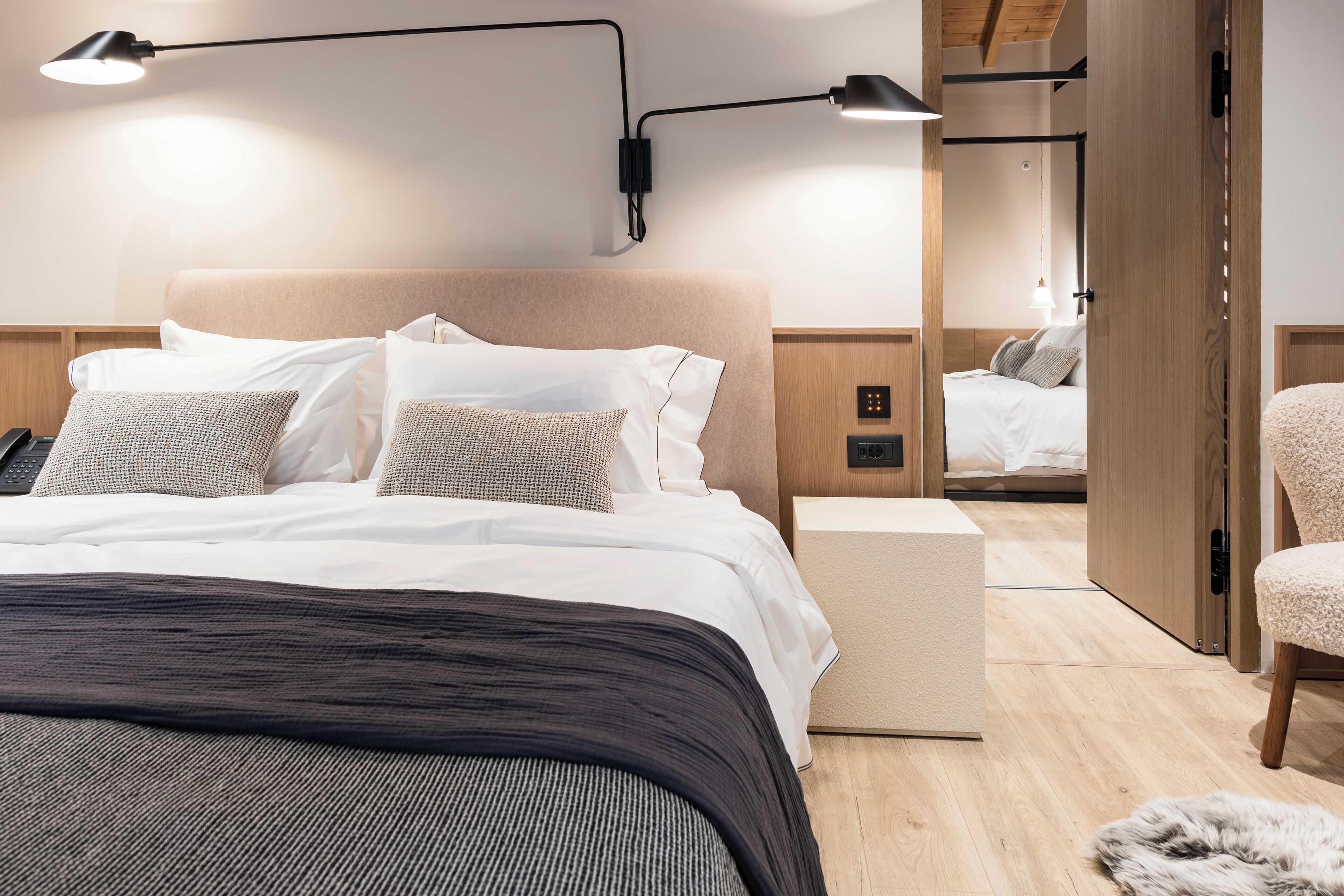 A modern hotel room with a large bed, bedside lamps, a wooden headboard, a nightstand, and a fluffy rug.