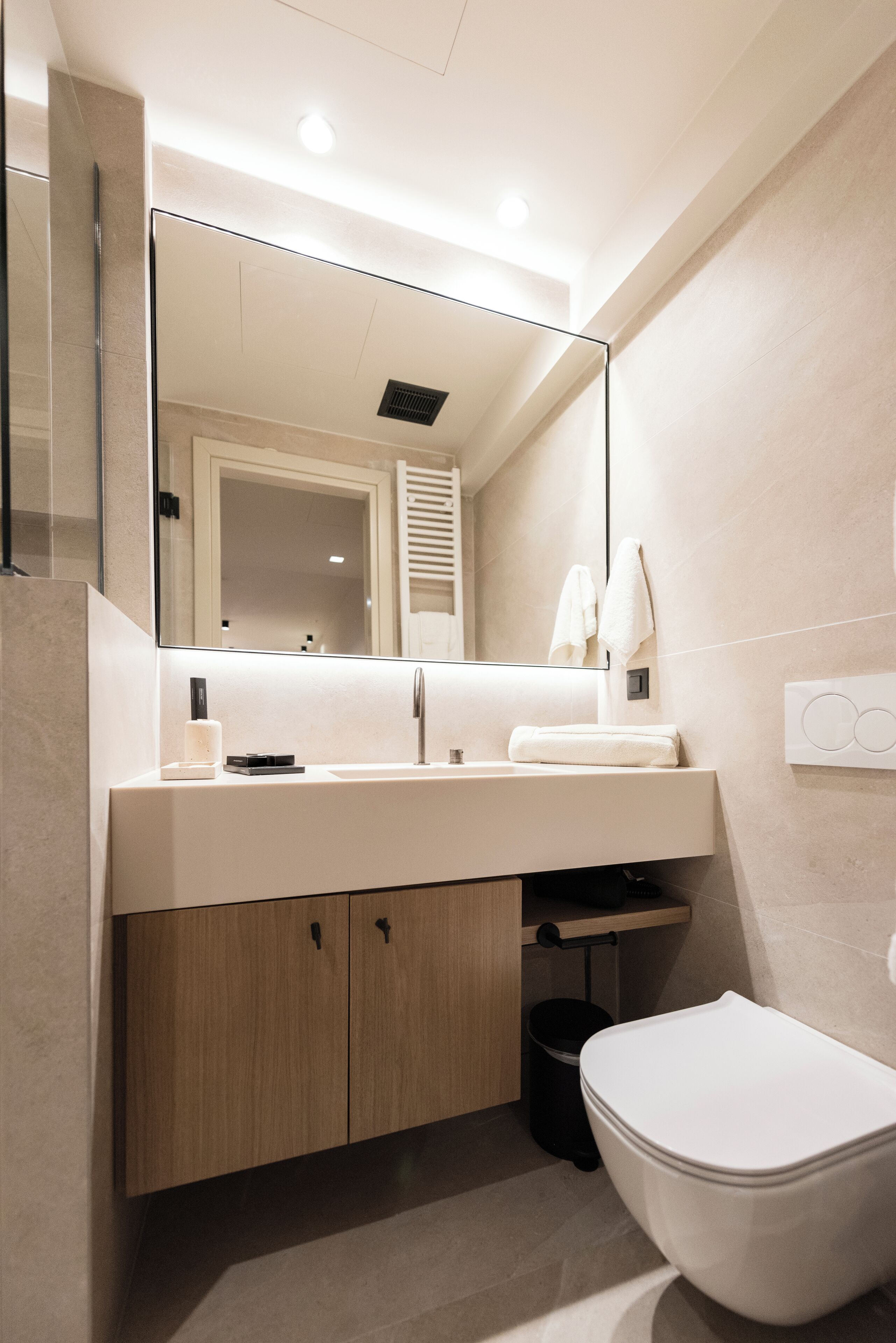 Une salle de bain moderne avec un grand miroir, un lavabo blanc, des meubles en bois, des toilettes et un porte-serviettes.