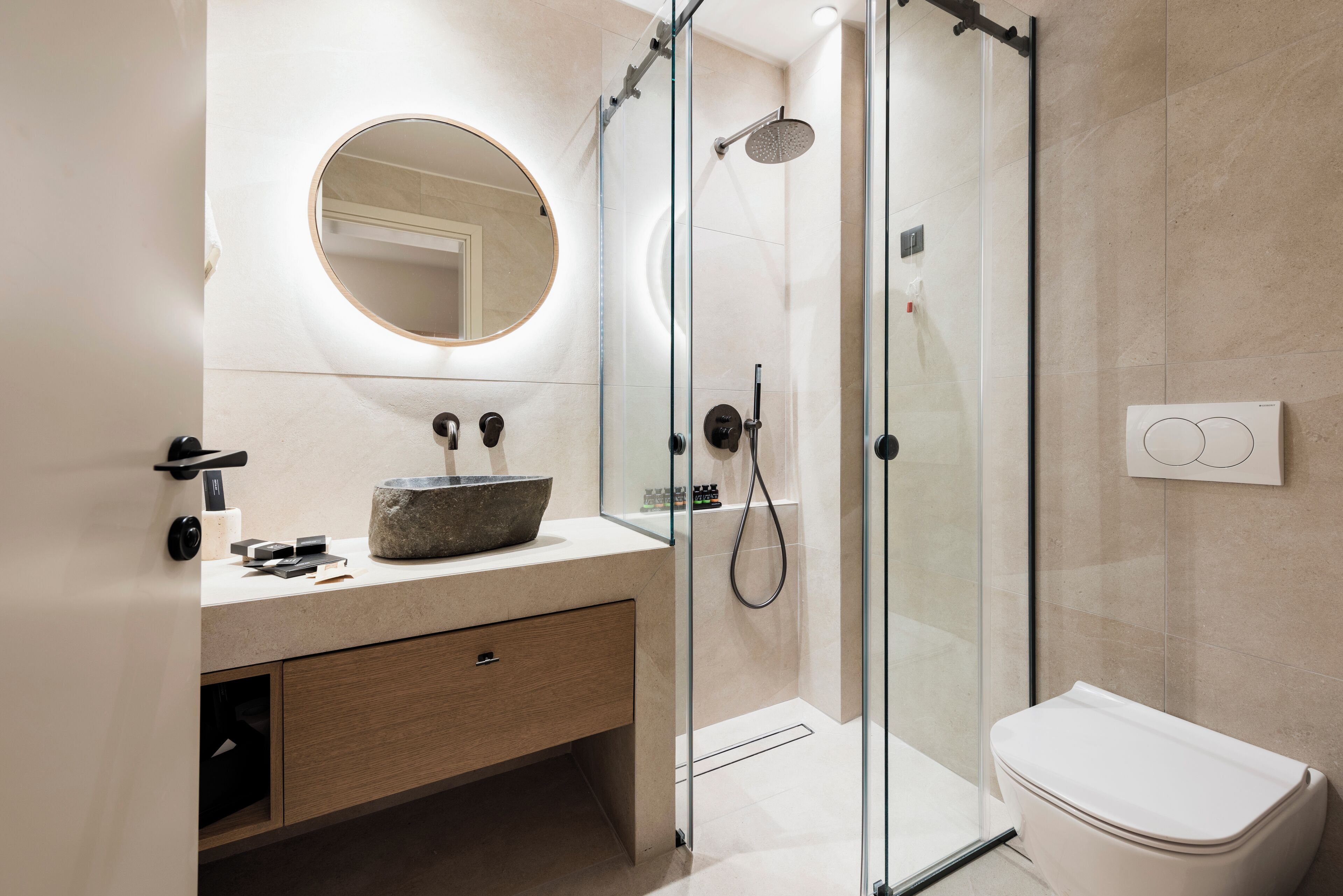 A modern bathroom with a glass-enclosed shower, a round mirror, a stone sink, and a toilet.
