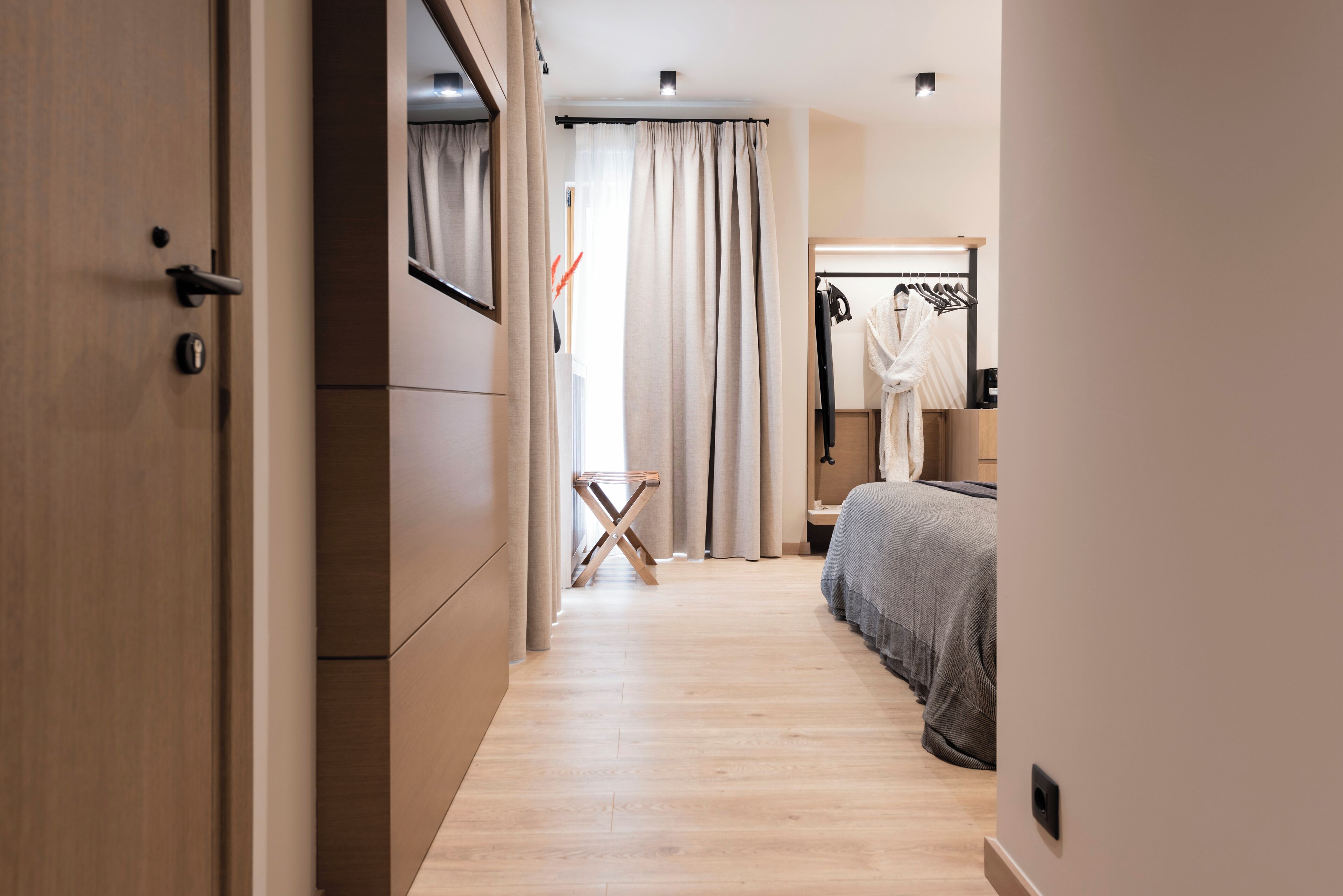 Una habitación de hotel moderna con suelo de madera, una cama con manta gris, un armario, un espejo y una ventana con cortinas.