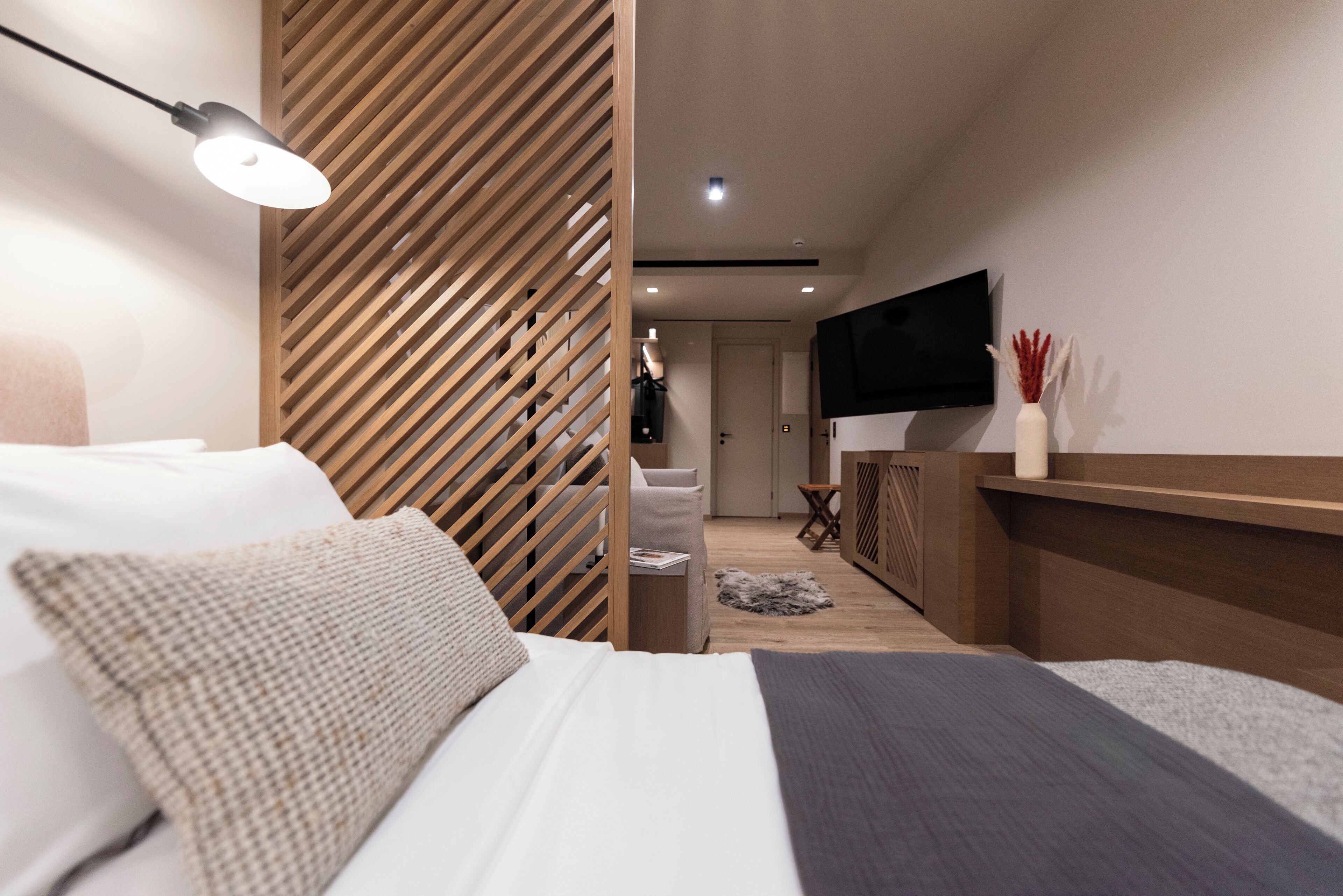 Quarto moderno com cama, luminária de cabeceira, TV fixada na parede e divisória de madeira.