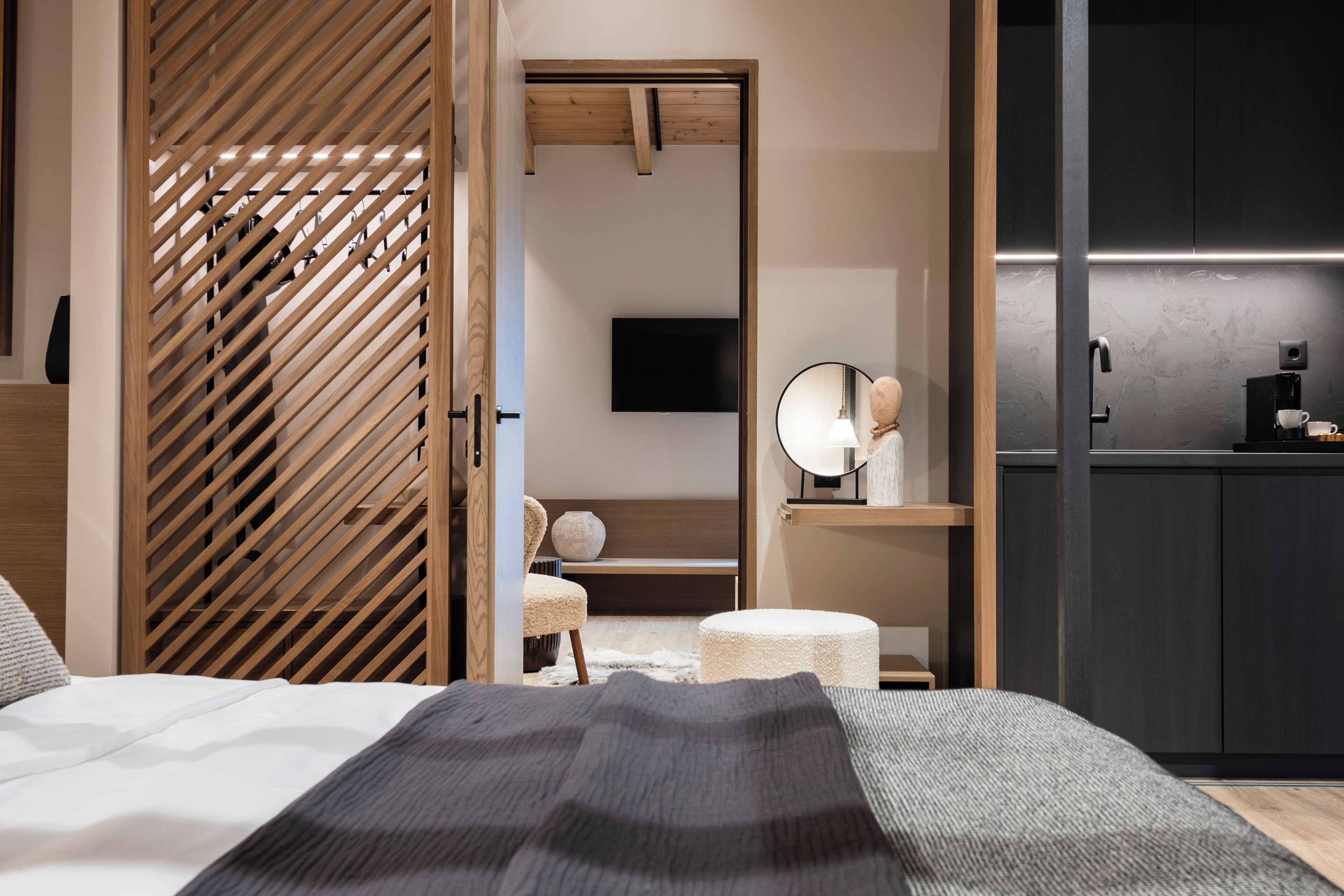A modern bedroom with a bed, a wooden slatted door, a small round table, a white chair, a television, and a coffee machine.