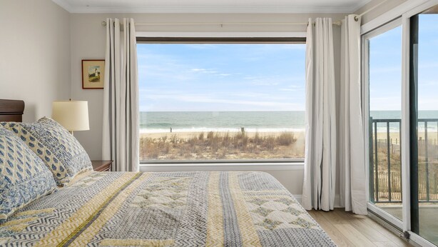 Ein Schlafzimmer mit großem Fenster mit Blick auf einen Strand, einem Bett mit gemusterten Betttextilien, einer Nachttischlampe und einem gerahmten Bild an der Wand.