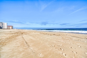 A beach with footprints leading to the water, a clear sky, and buildings in the background.
