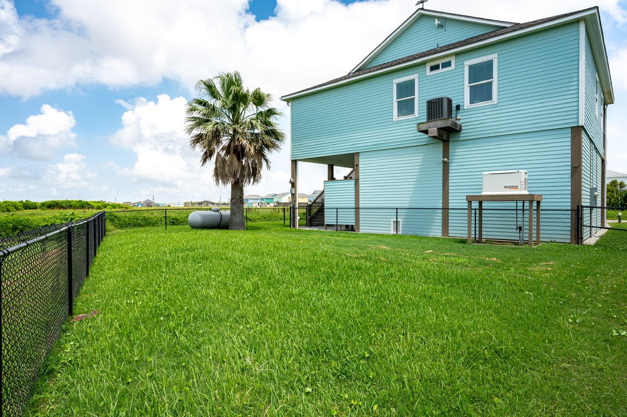 A light blue house with a fenced yard, a palm tree, and a generator on the porch.