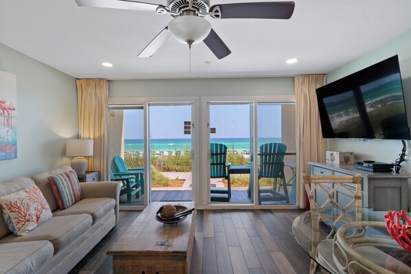 A living room with a view of the beach, a ceiling fan, and a flat-screen TV.
