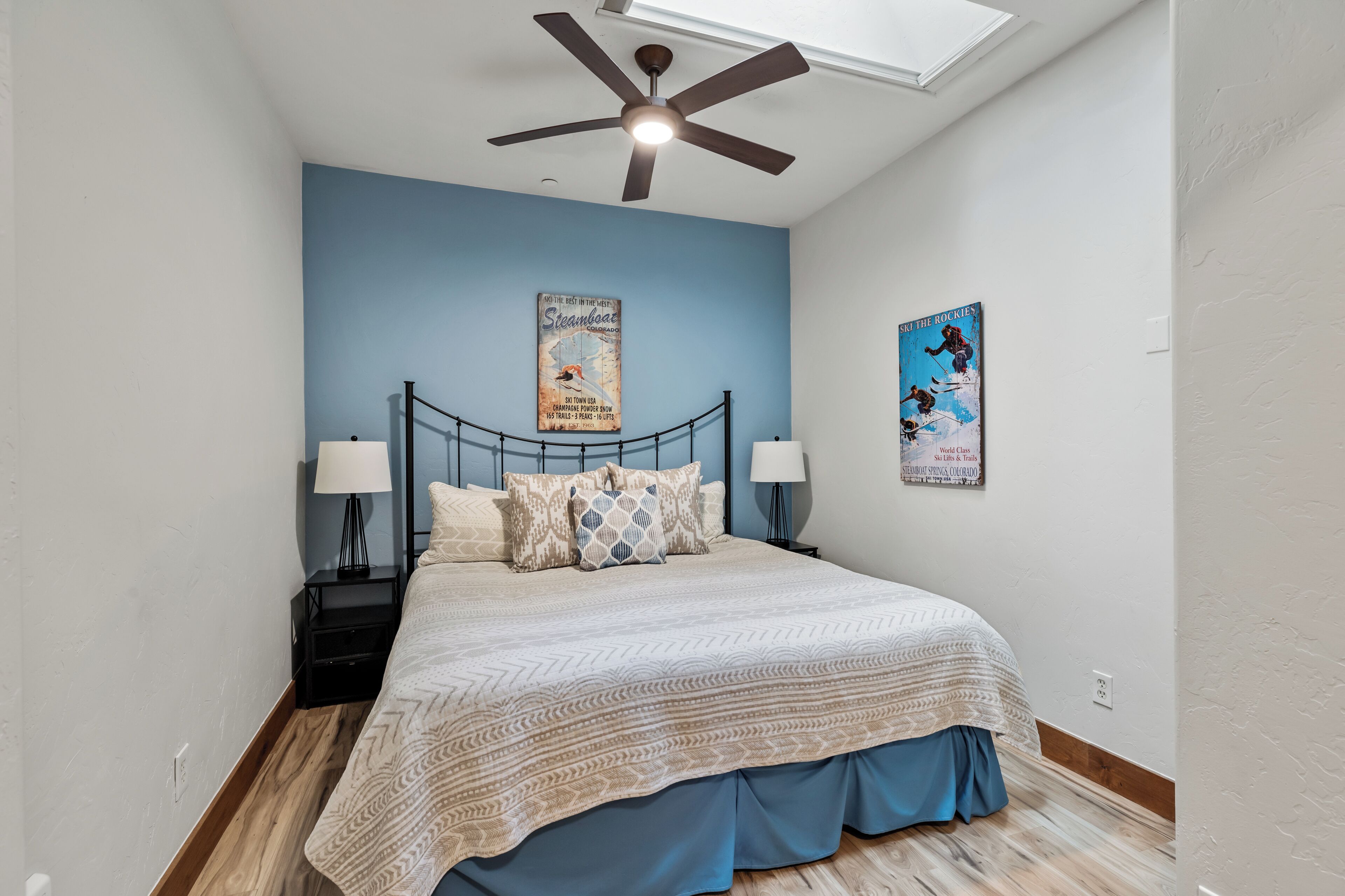 Un dormitorio con una cama, dos mesitas de noche, un ventilador de techo y dos carteles en la pared.