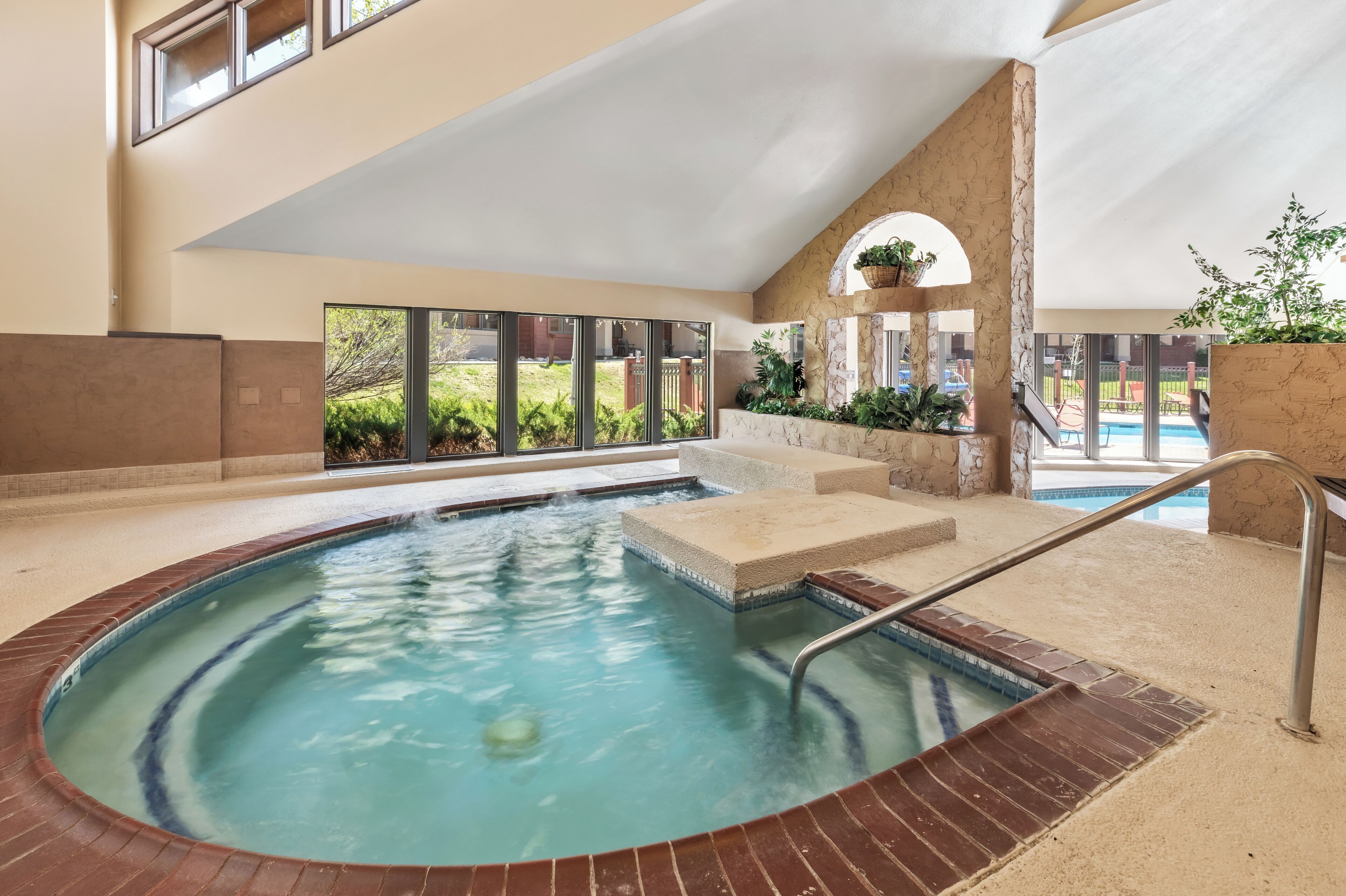 Ein Whirlpool mit Backsteinrand, einer Metallleiter und Blick auf einen Garten mit Schwimmbad und Grünflächen.
