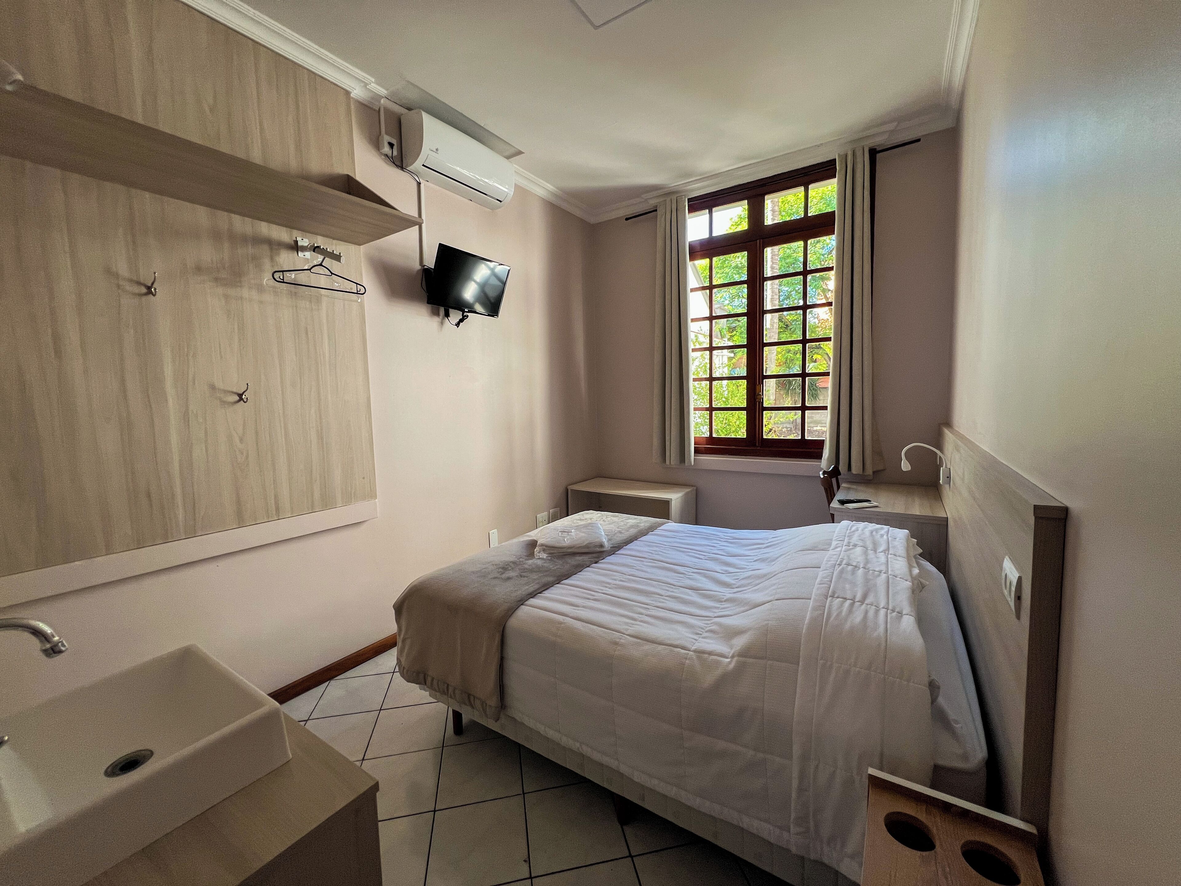A hotel room with a bed, a TV mounted on the wall, a window with curtains, a bedside table, and a bathroom with a sink and mirror.