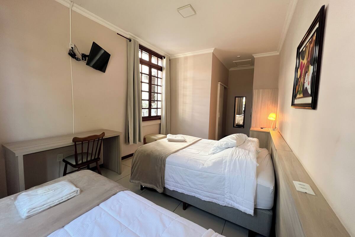 A hotel room with two beds, a desk, a chair, a TV, a window with curtains, a bedside lamp, and a framed picture on the wall.