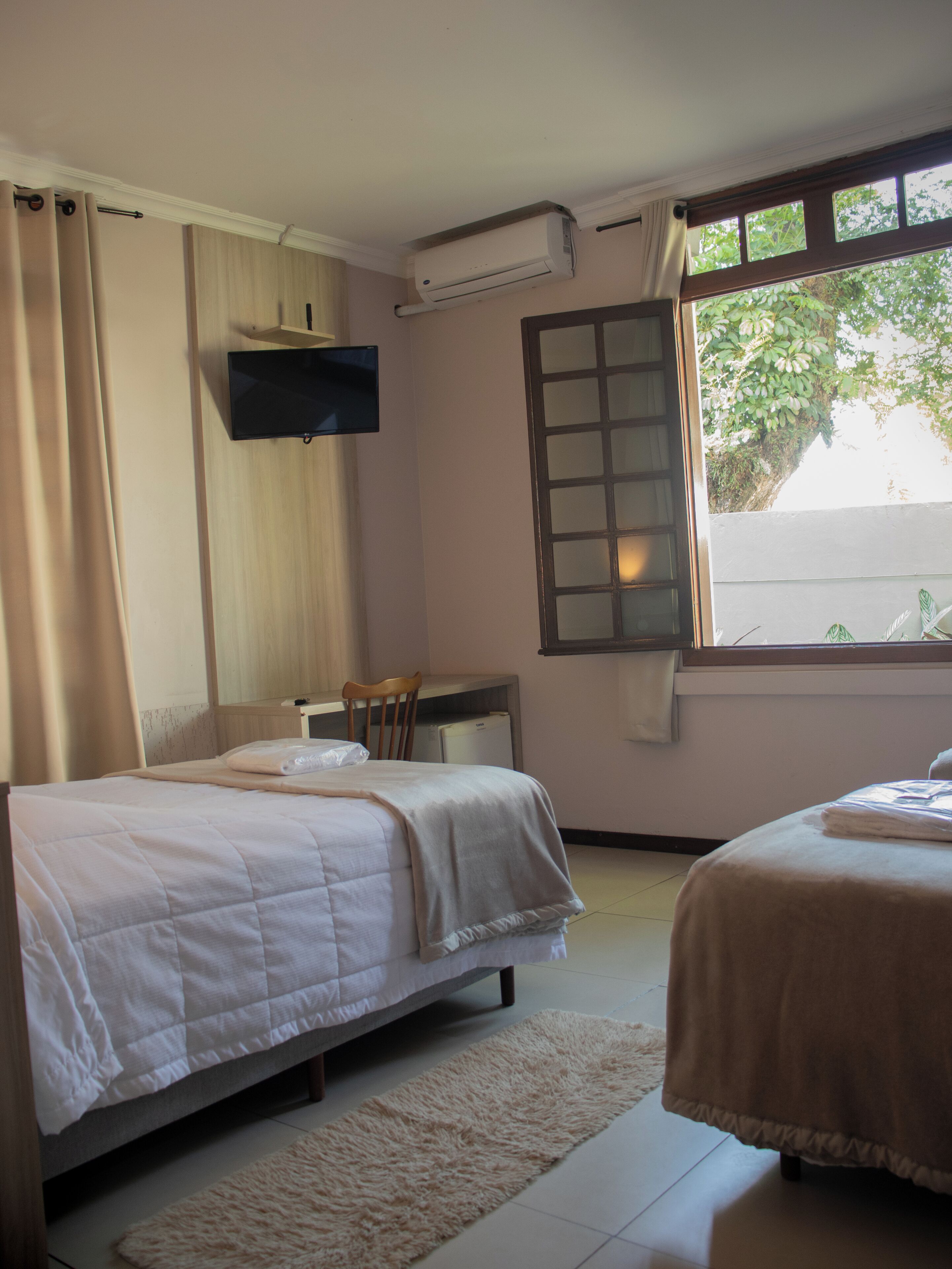 Een slaapkamer met twee bedden, een bureau, een stoel, een airconditioning en een raam met uitzicht op groen.