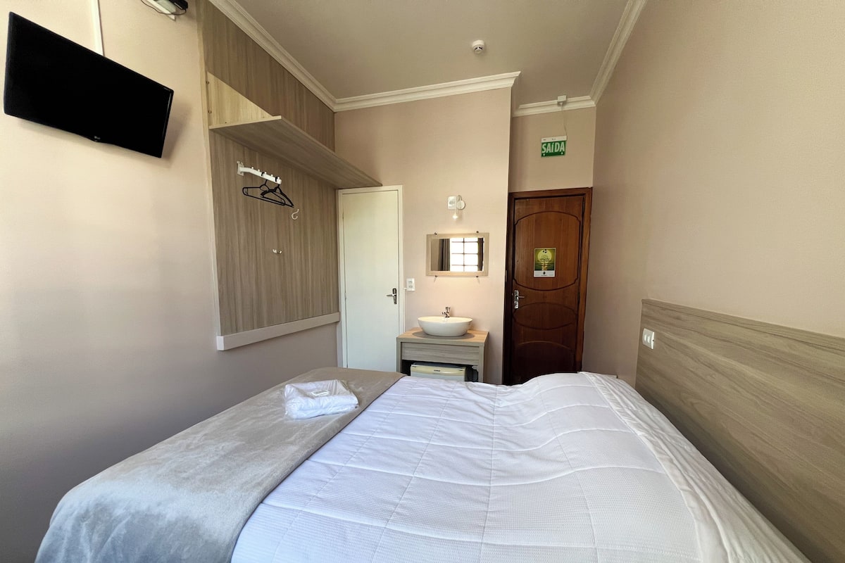 A hotel room with a bed, a bedside table with a sink, a mirror, and a door with a sign.