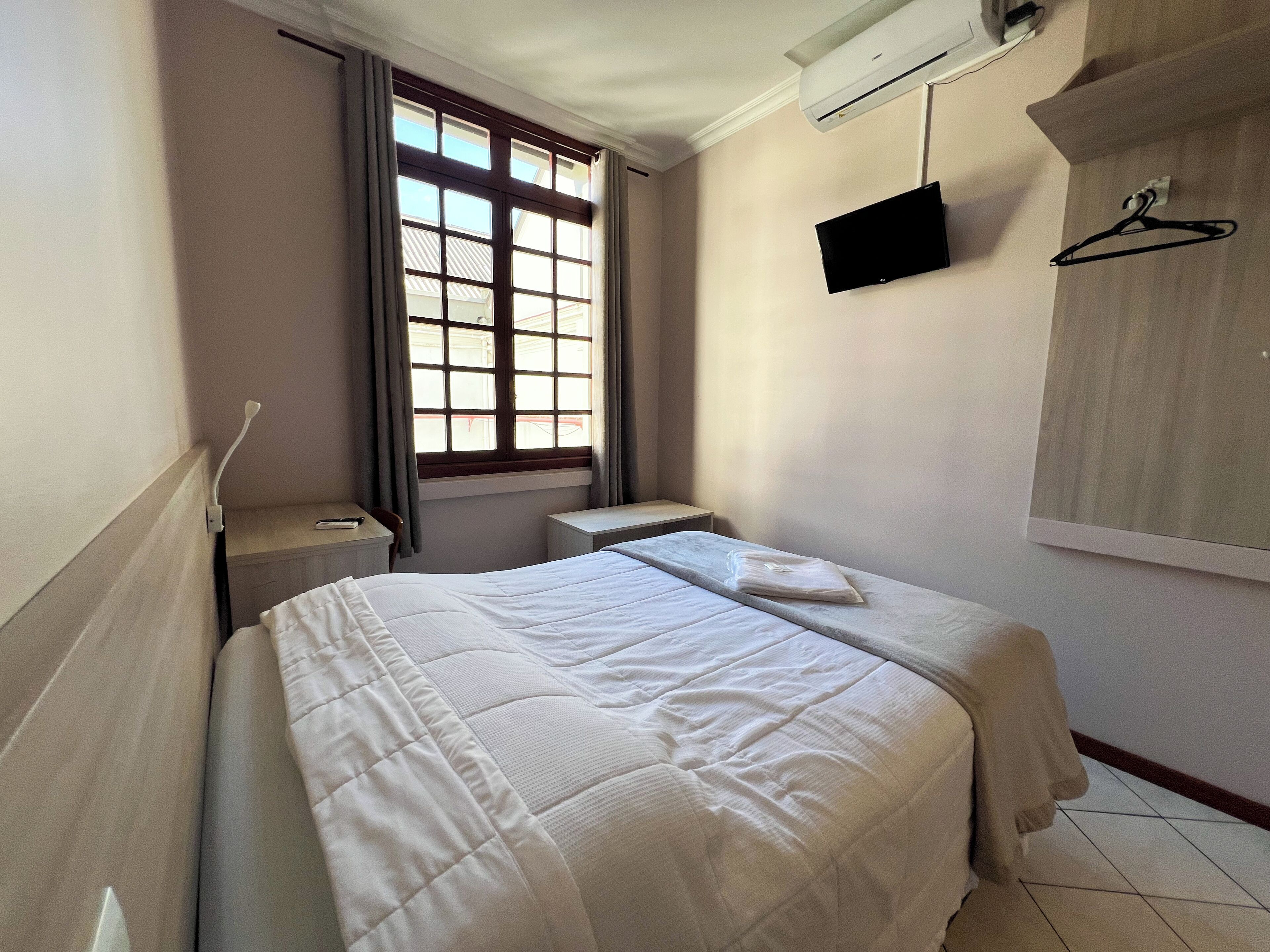 Una cama bien hecha en una habitación de hotel, con una ventana, un televisor de pantalla plana montado en la pared y una mesita de noche.