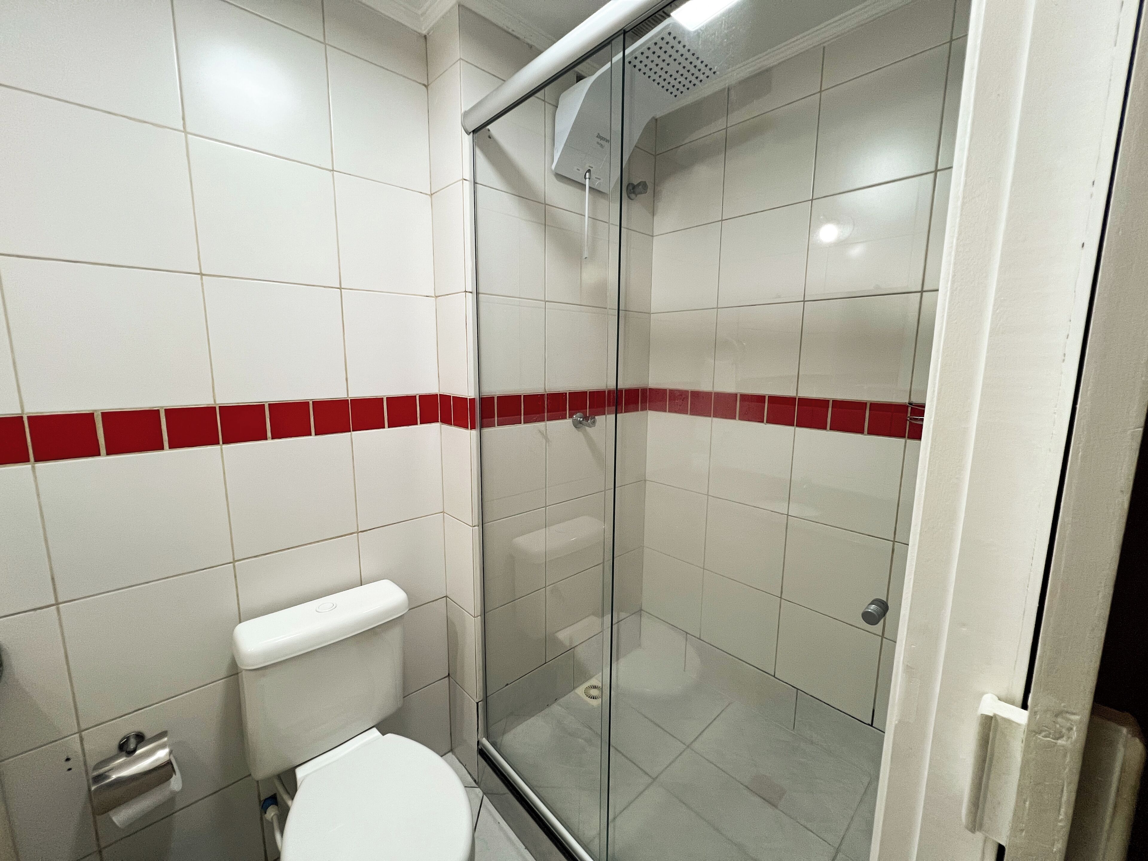 A bathroom with white tiles, a glass shower enclosure, and a red tile accent strip.