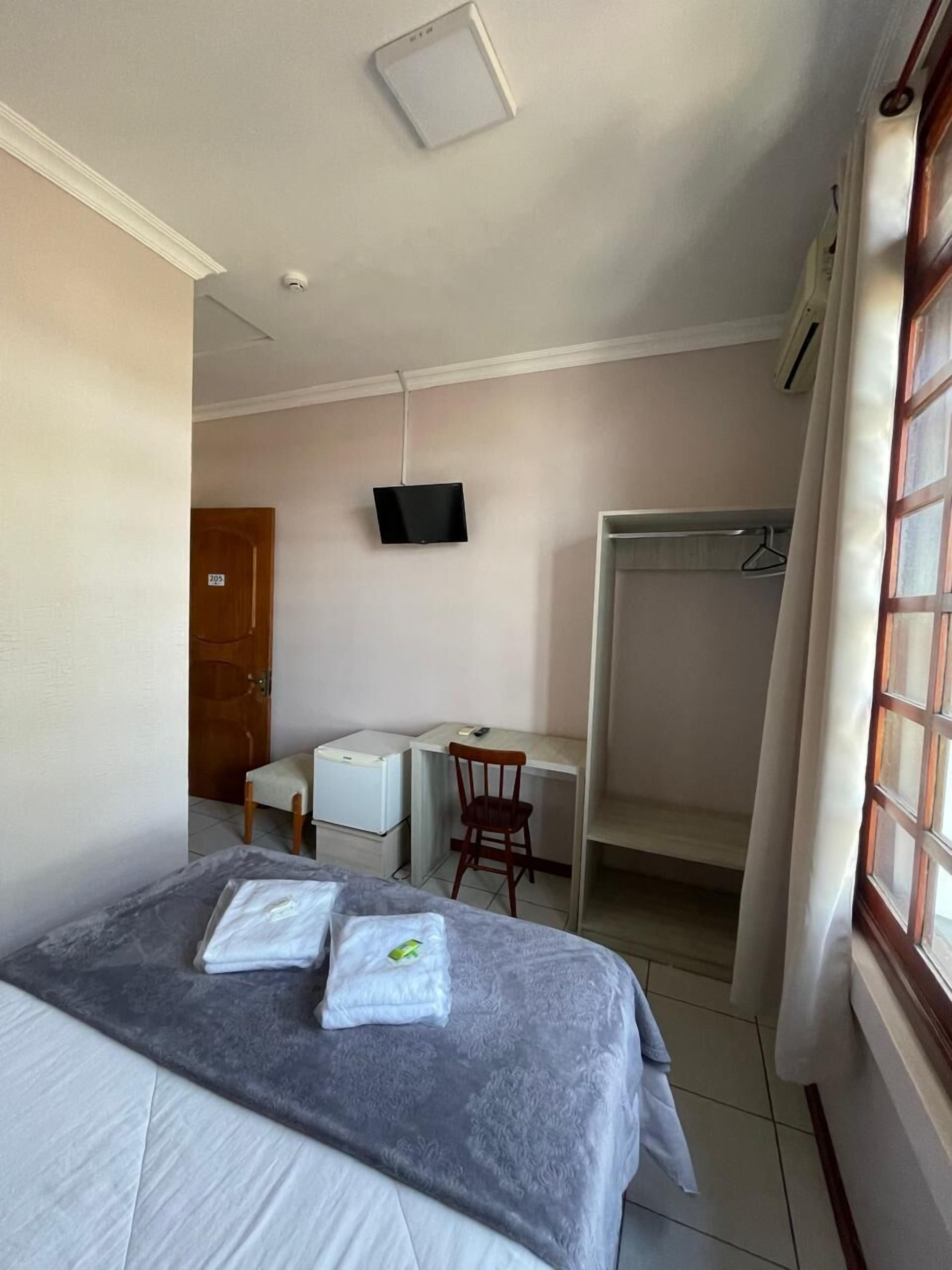 Quarto de hotel com cama, escrivaninha, cadeira, televisão e guarda-roupa.