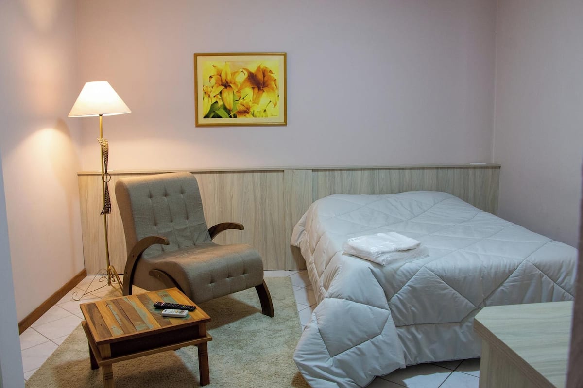 A hotel room with a bed, a chair, a table with remote controls, a lamp, and a painting on the wall.