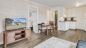 A living room with a TV, wooden furniture, a dining table, and a kitchen area with white cabinets and appliances. - Flirtly Flamingo Rp203a 2 Bedroom Duplex by RedAwning (Rockport)