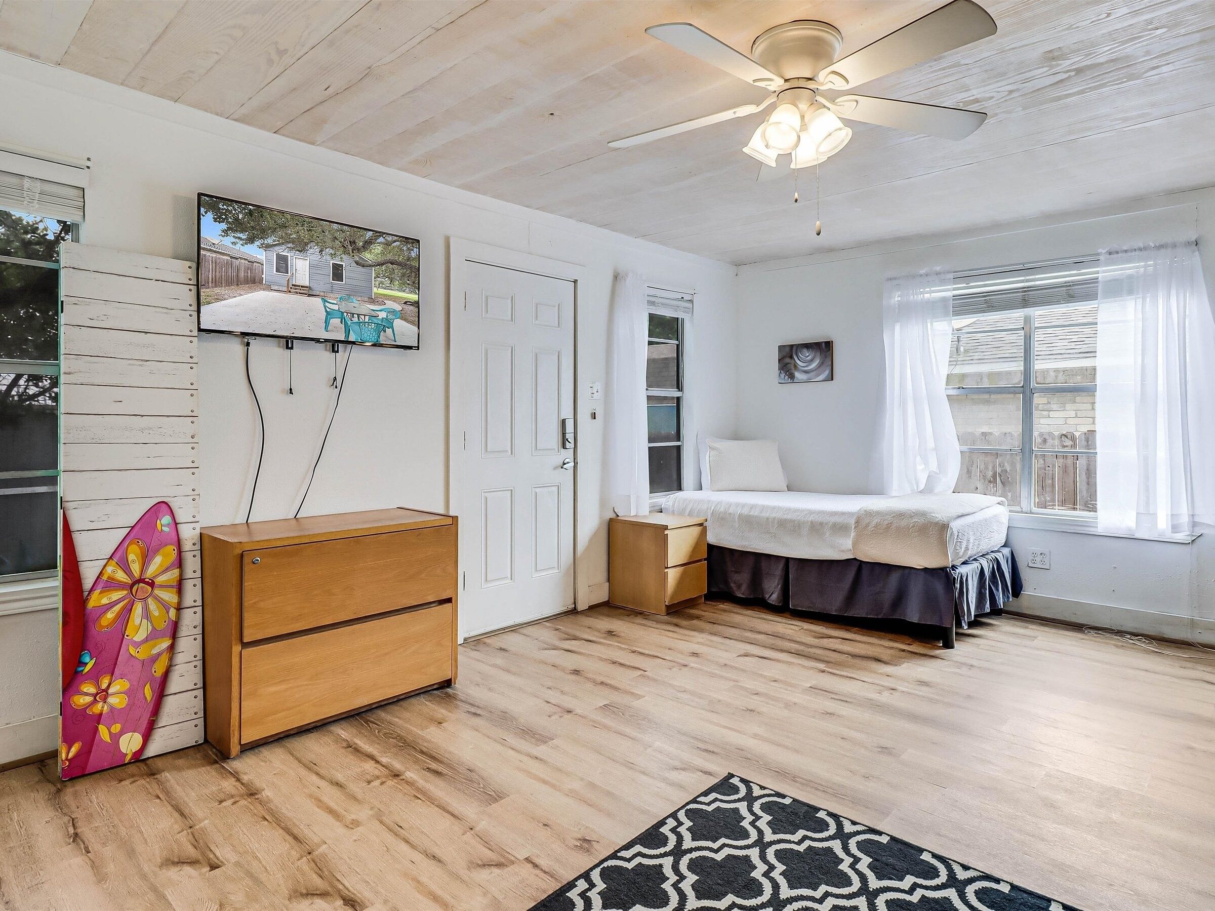 Une chambre à coucher avec un lit, un ventilateur de plafond, un téléviseur fixé au mur et une fenêtre avec des rideaux.