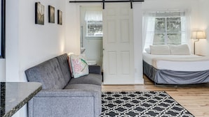 A room with a grey sofa, a bed with a grey bed skirt, a black and white patterned rug, and a window with a white curtain. - Merry Macaw Rp203b 1 Bedroom Duplex by RedAwning (Rockport)
