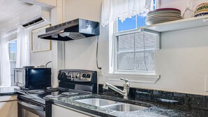 A kitchen with a stove, oven, and sink. There is a window with blinds and a shelf with plates. - Merry Macaw Rp203b 1 Bedroom Duplex by RedAwning (Rockport)