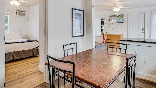 A room with a wooden floor, a bed with white bedding, a wooden dining table with chairs, and a kitchen area with a counter and chairs. - Merry Macaw Rp203b 1 Bedroom Duplex by RedAwning (Rockport)