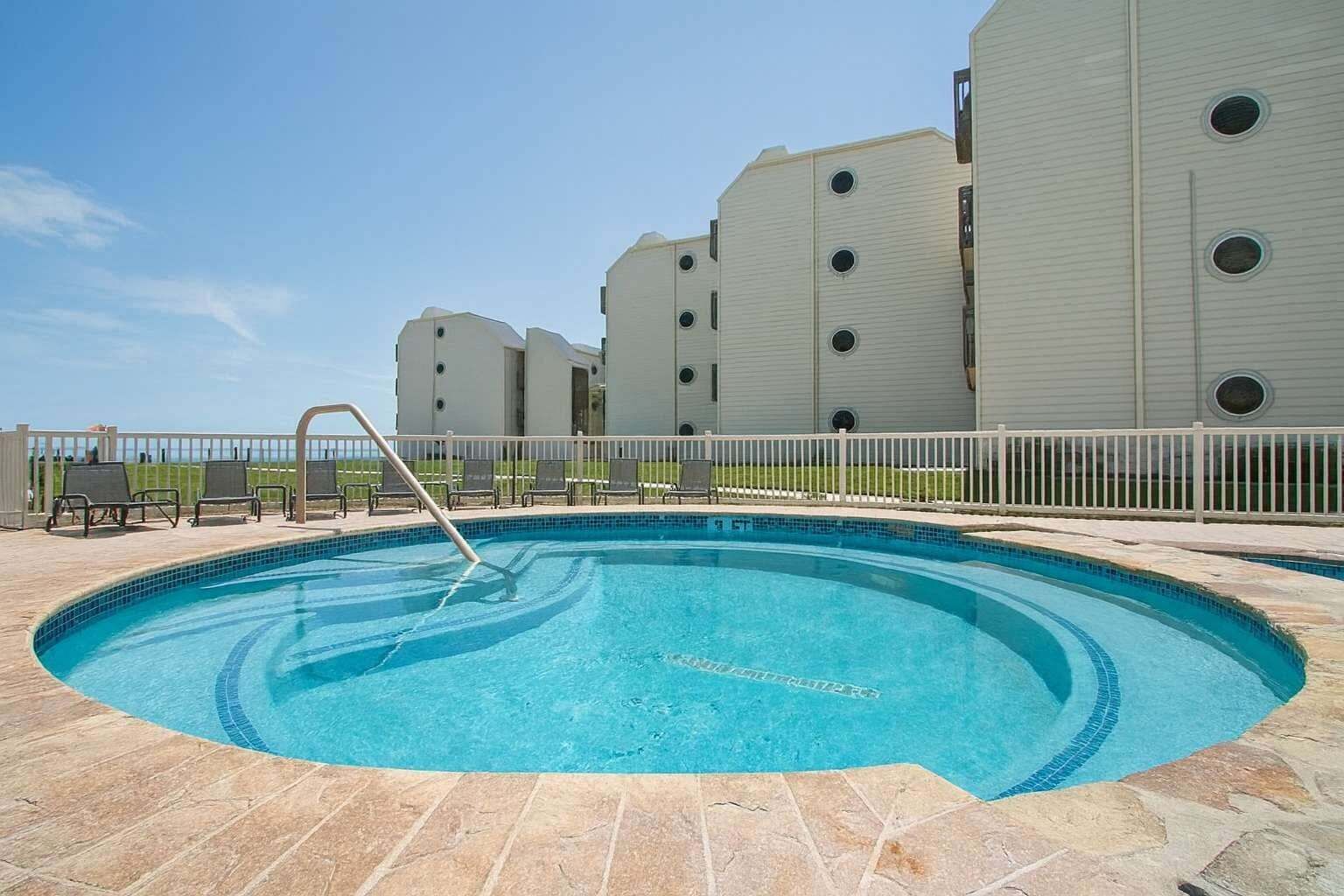 Una piscina con borde de azulejos, rodeada por un suelo de piedra. Se observan sillas de descanso y una cerca blanca al fondo.