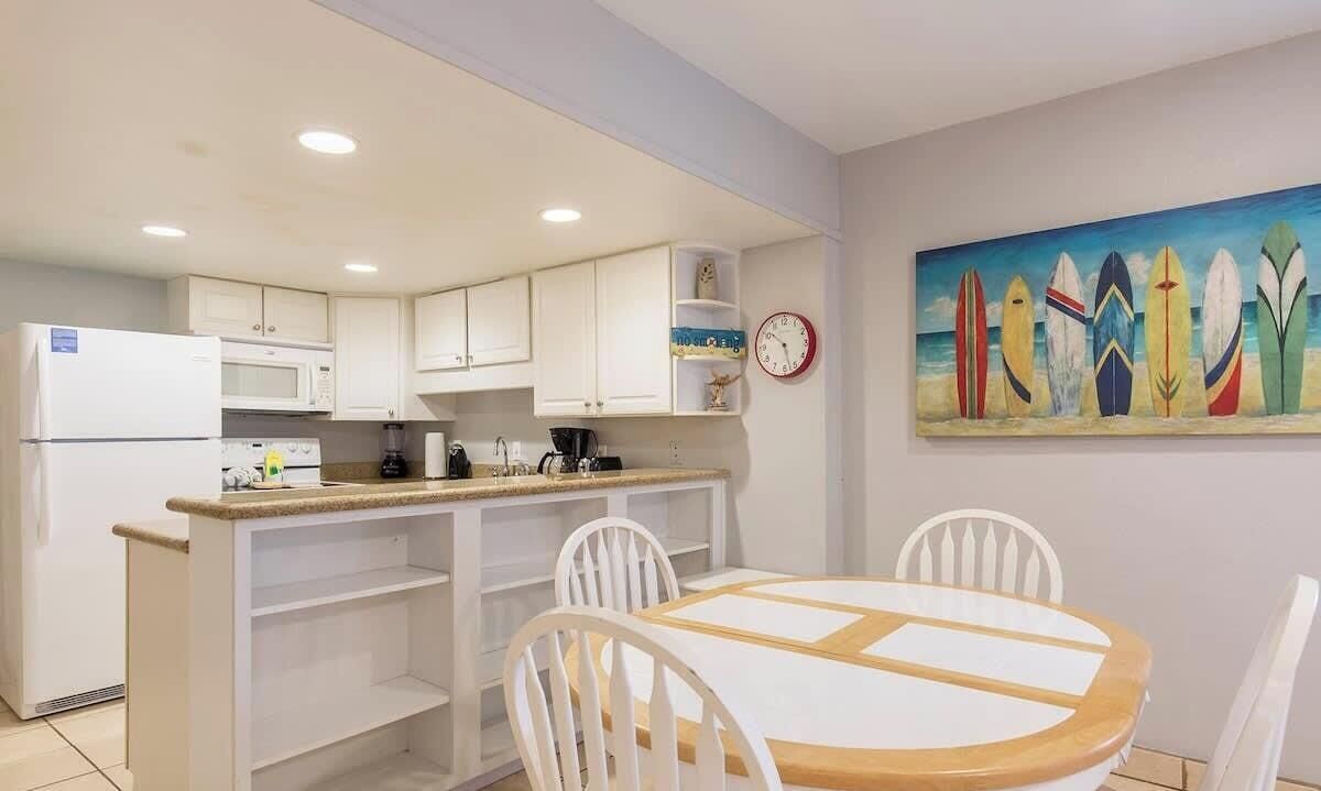 Een compacte keuken met witte kastjes, een magnetron en een spoelbak. Er is een eethoek met een ronde tafel en witte stoelen. Een klok hangt aan de muur en boven de tafel hangt een kleurrijk schilderij van een surfboard.