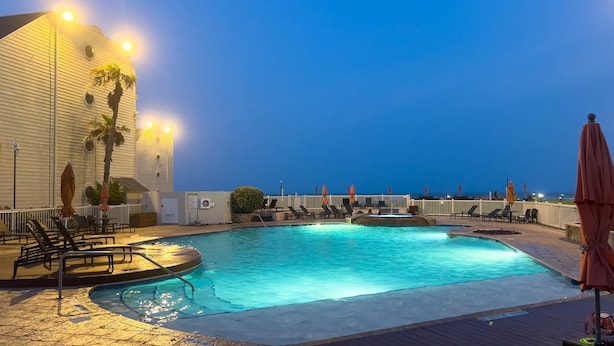 Ein Poolbereich mit Liegestühlen und Sonnenschirmen, ein Holzdeck sowie ein Gebäude mit beleuchteter Fassade.