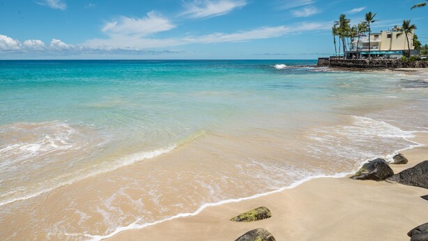 A beach with clear turquoise water, a sandy shore, and a building with a balcony and palm trees in the background.