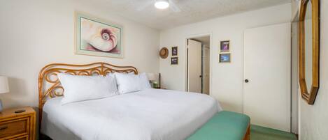 Un dormitorio con cama, mesita de noche, banco verde, ventilador de techo y una puerta.