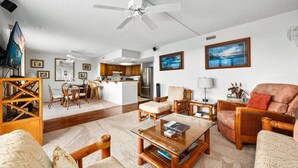A spacious living room with a glass coffee table, wooden furniture, a ceiling fan, and a kitchen in the background.