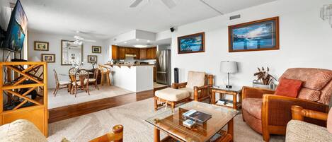 A spacious living room with a glass coffee table, wooden furniture, a ceiling fan, and a kitchen in the background.
