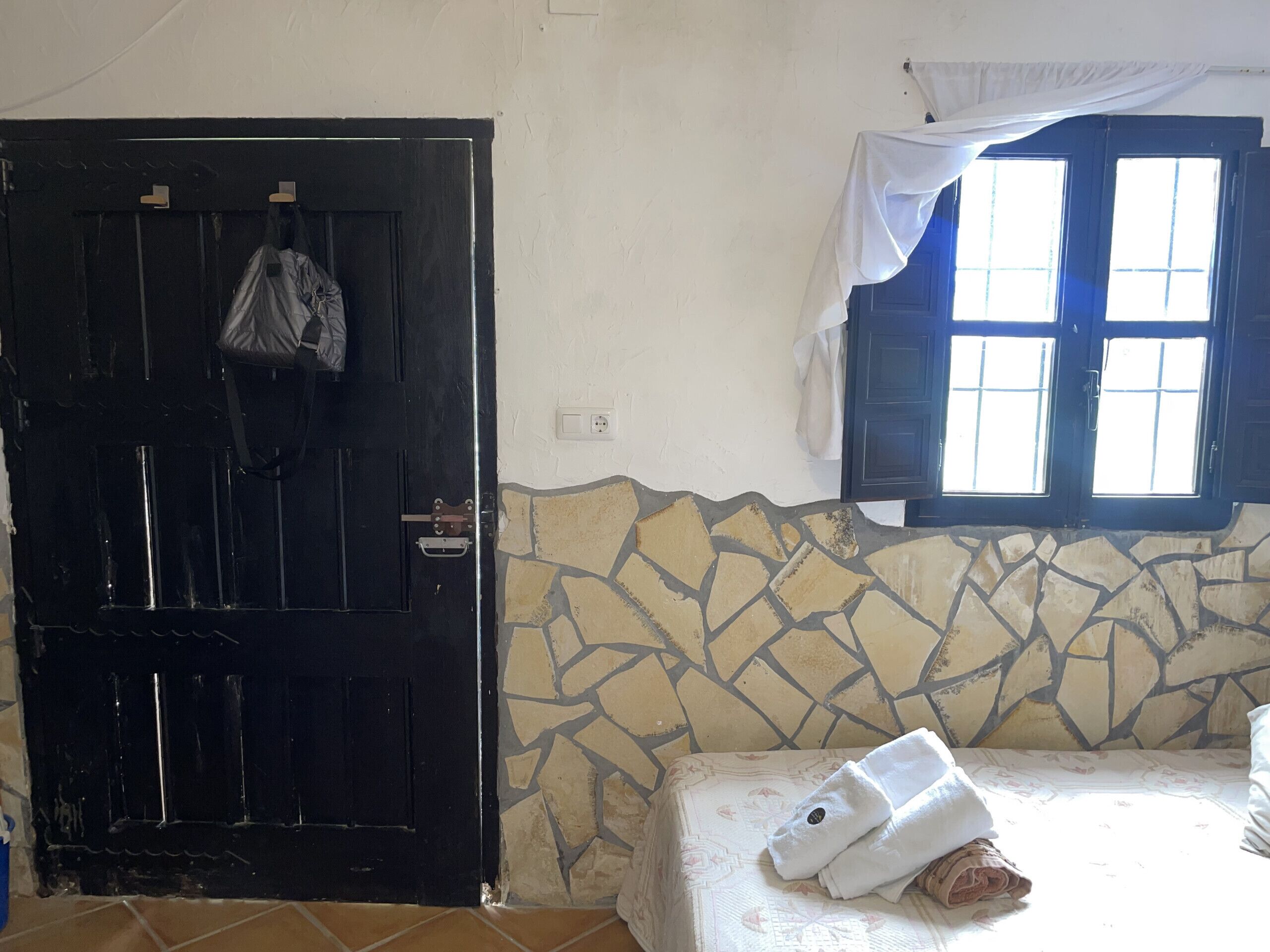 Une pièce avec une porte noire, une fenêtre ornée de rideaux blancs, un lit recouvert d’une couvre-lit à motifs et un mur en pierre.