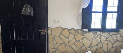 Une pièce avec une porte noire, une fenêtre ornée de rideaux blancs, un lit recouvert d’une couvre-lit à motifs et un mur en pierre.
