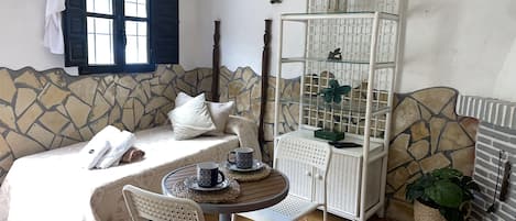 A cozy room with a bed, a small table with chairs, a white cabinet with glass shelves, and a stone-patterned wall.