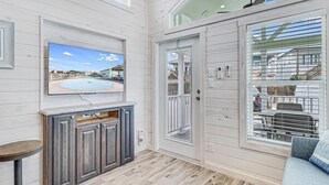 Ein Zimmer mit einem großen Fernseher, der eine tropische Strandlandschaft zeigt, einem Holzboden, einem grauen Schrank und einer Glastür, die nach draußen führt.