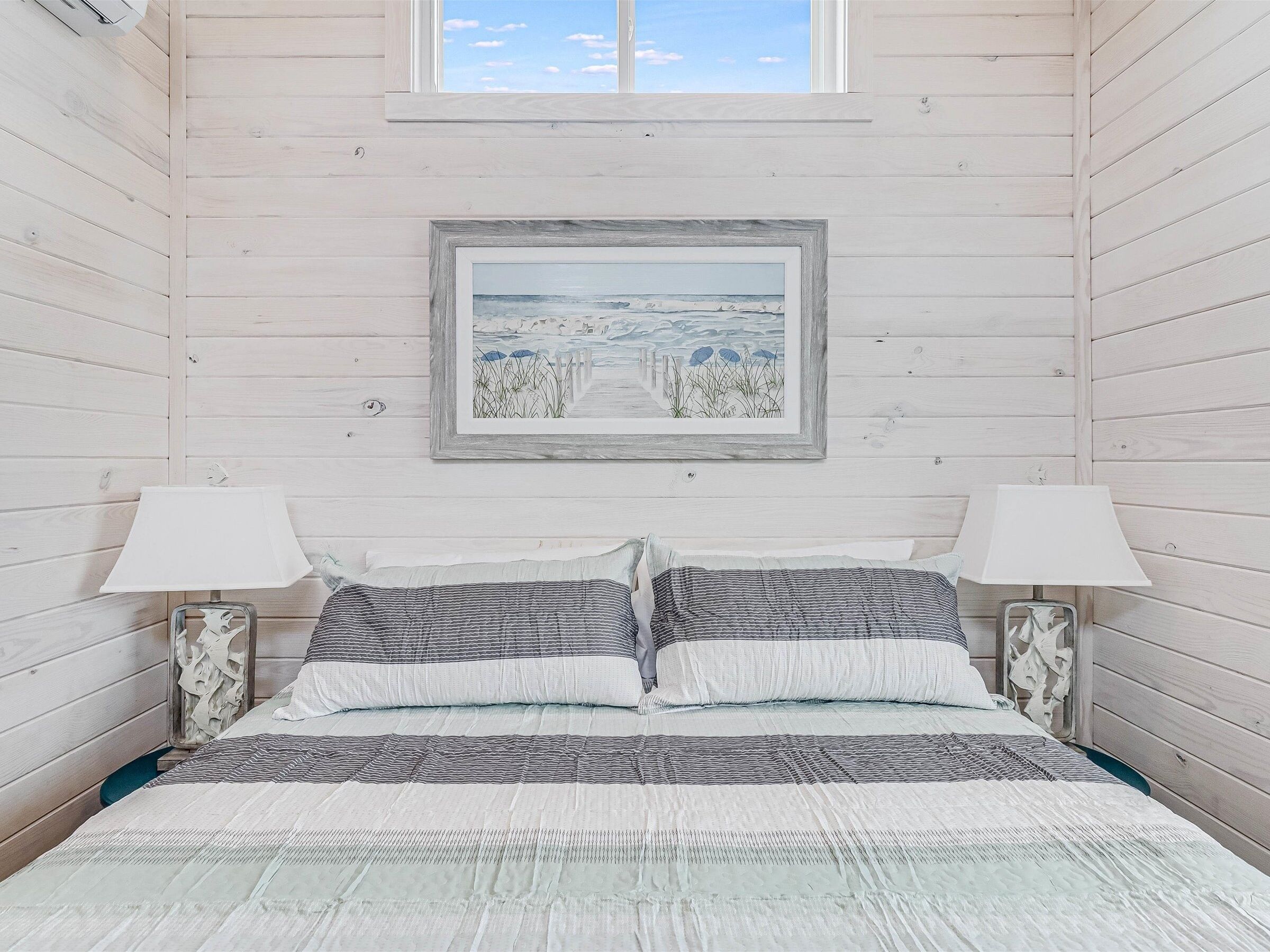 Un dormitorio con pared de madera, un cuadro enmarco con una escena de playa, dos lámparas de noche y una cama con ropa de cama a rayas.