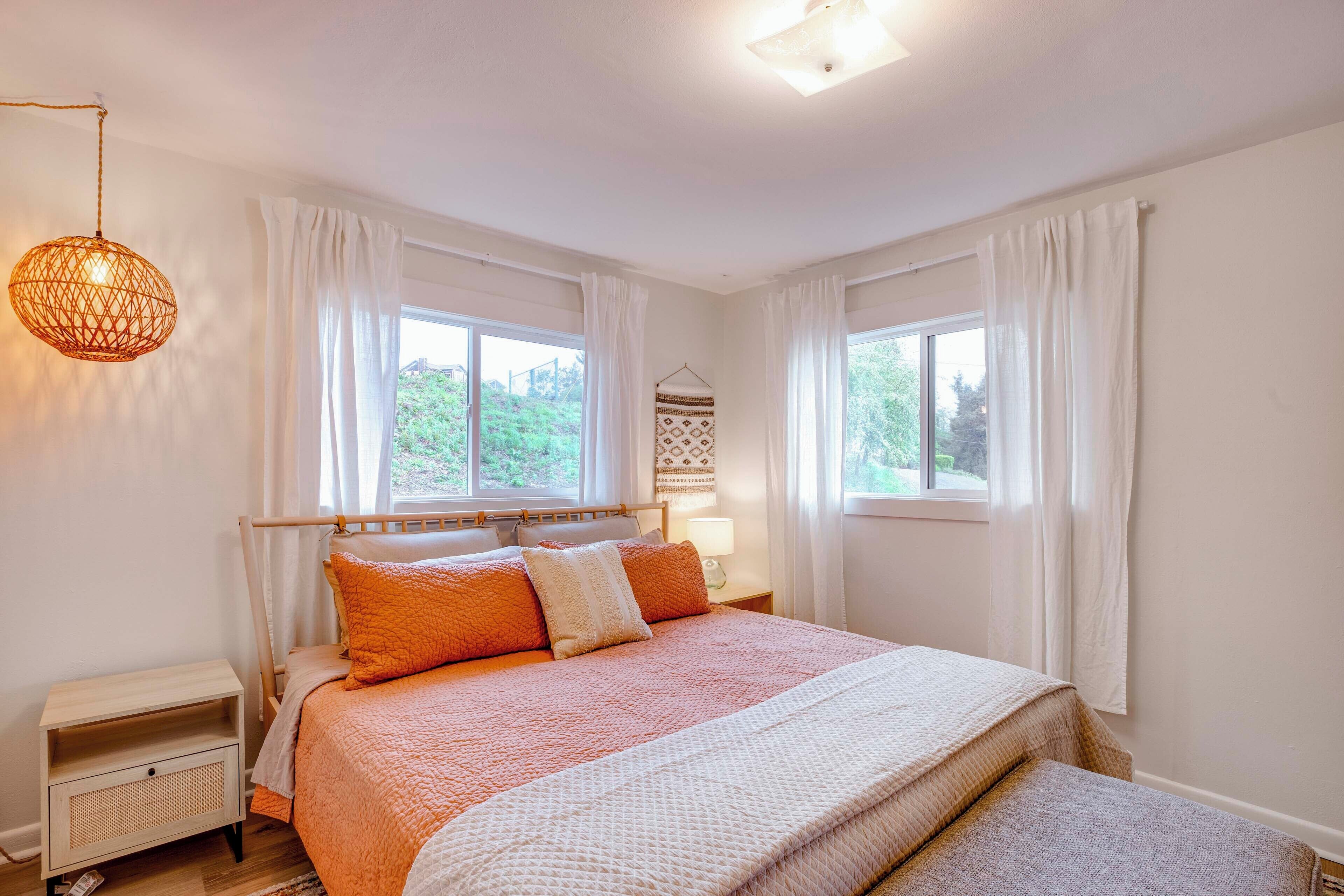Ein Schlafzimmer mit einem Bett, Nachttischen, einer Pendelleuchte und zwei mit Vorhängen versehenen Fenstern.