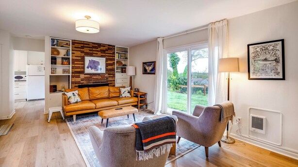 A modern living room with a leather sofa, a grey armchair, a coffee table, and a bookshelf. There is a large window with a view of the outdoors. - Bay City Retreat ~ 1/2 Mi to Local Beach! (Bay City)