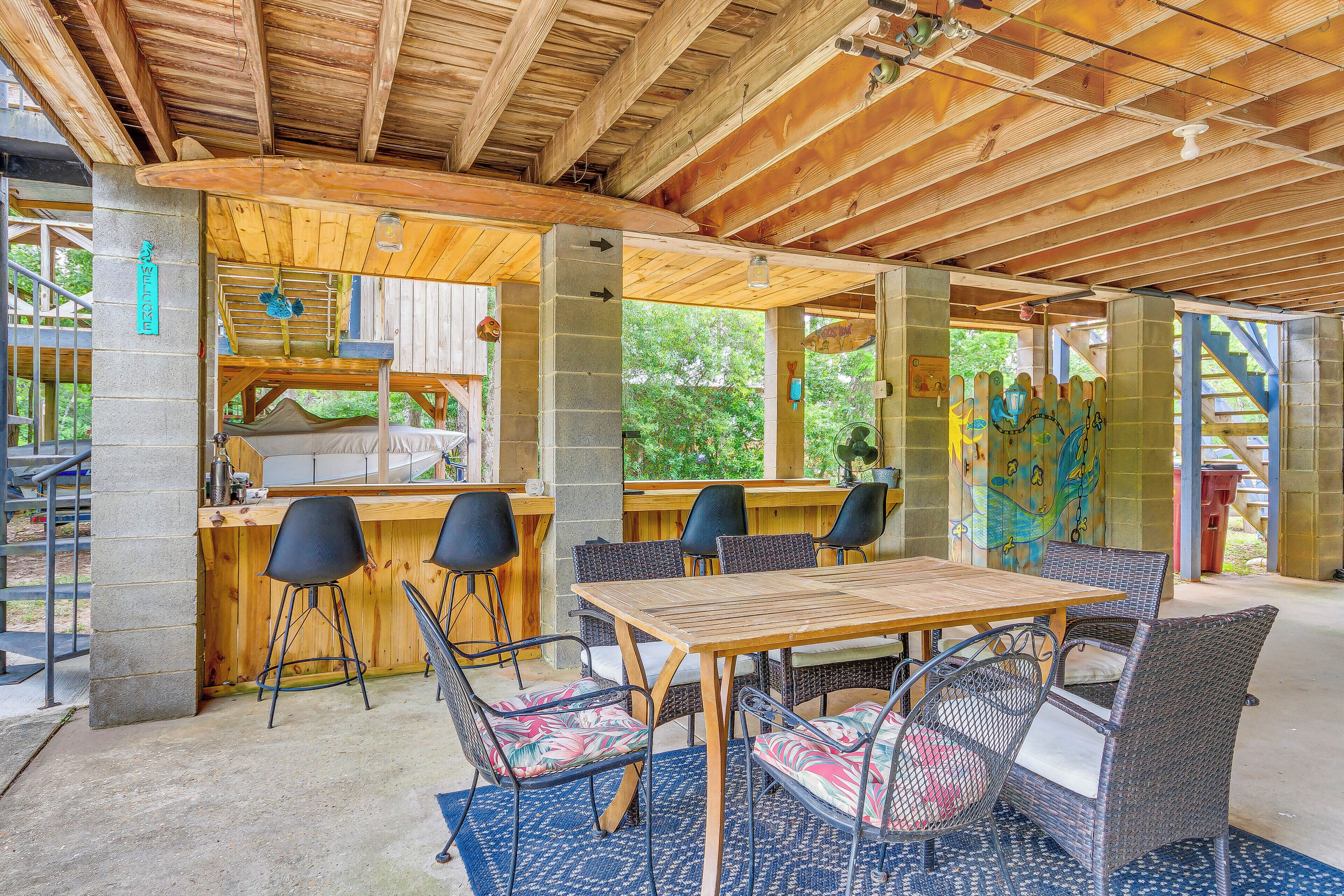 An outdoor patio with a wooden table and wicker chairs, a bar area with stools, and a view of greenery.
