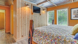 Une chambre avec un mur en bois, un lit recouvert d’une couvre-lit à motifs floraux, un téléviseur fixé au mur et une fenêtre avec des stores.
